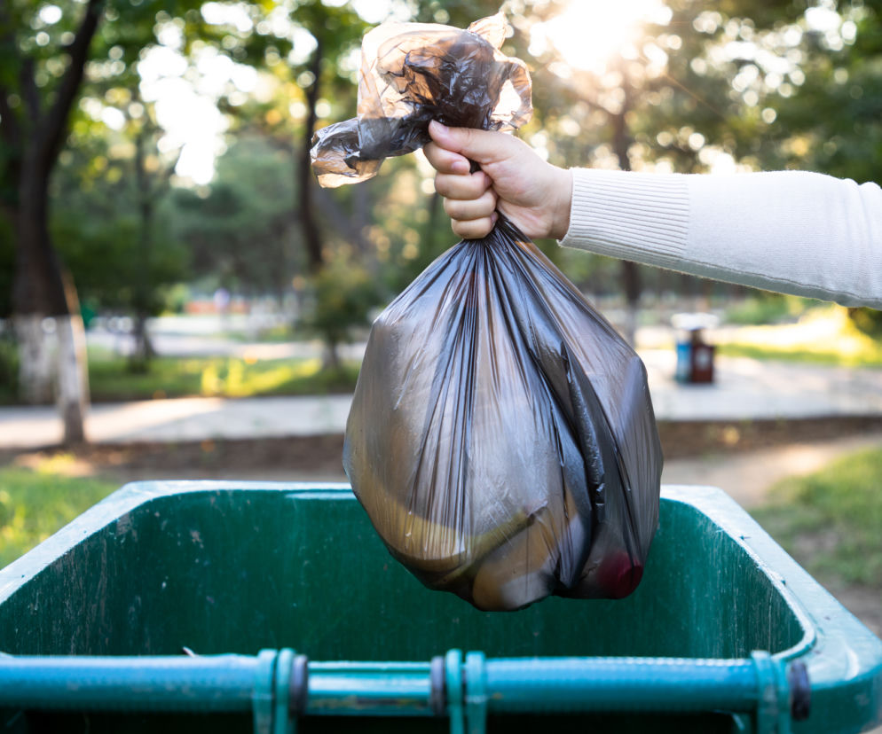 Osoba wyrzucająca czarny worek na śmieci do zielonego kontenera w parku. Fotografia symbolizuje konieczność segregacji i opłat za odpady, o których przeczytasz więcej na Super Biznes.