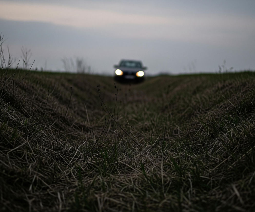 Ciemny samochód z włączonymi, jasnymi światłami przednimi zbliża się z oddali, widoczny w centralnej części kadru, powyżej linii horyzontu. Przed nim rozciąga się pofalowane pole, na którym dominują sucha trawa i resztki roślin, tworząc wyraźne, ciemne bruzdy prowadzące w głąb kadru. Jasnoszare niebo zajmuje górną część obrazu, delikatnie rozświetlając tło za oddalonym samochodem.