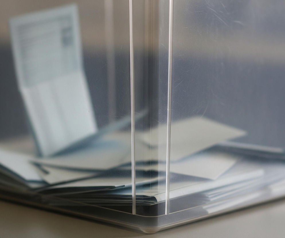 Przezroczysta urna z pleksi wypełniona jest licznymi, jasnoniebieskimi kartkami papieru, stanowiącymi główny obiekt na zdjęciu. Kartki leżą chaotycznie jedna na drugiej, tworząc stos, a jedna z nich jest widoczna wyraźnie, wystając pionowo w lewym górnym rogu. Cała urna stoi na jasnym, matowym blacie, a tło za nią jest rozmyte, ciemnoszare, z widocznymi jaśniejszymi refleksami.