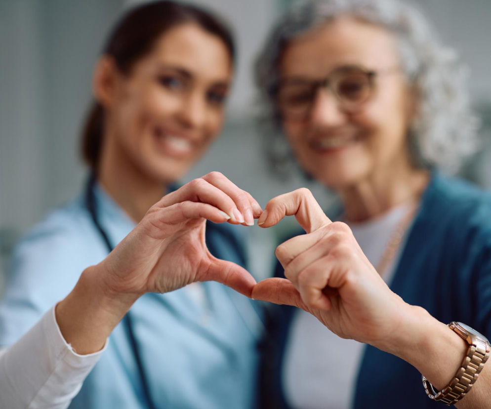 Kobiety, młodsza lekarka i starsza pacjentka, łączą dłonie w symbol serca, symbolizując troskę o zdrowie serca po menopauzie. Obie uśmiechnięte, w tle rozmazane sylwetki, co podkreśla skupienie na geście. Więcej na ten temat przeczytasz na portalu Poradnik Zdrowie.