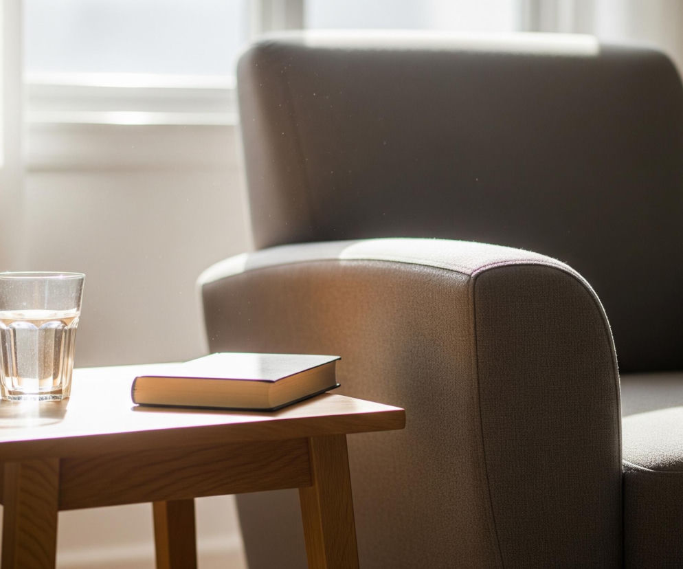 Centralnie na drewnianym, jasnobrązowym stoliku stoi szklanka wypełniona wodą oraz leży książka w ciemnej oprawie z jasnymi brzegami kartek. Stolik umiejscowiony jest obok szarego fotela, widocznego z prawej strony, z wyraźnie zaznaczoną fakturą materiału. W tle, po lewej stronie, rozświetlone światłem wpadającym przez okno, widoczne są jasne, lekko beżowe zasłony. Cała scena jest skąpana w naturalnym świetle, które tworzy wyraźne cienie i podkreśla drobinki kurzu unoszące się w powietrzu.