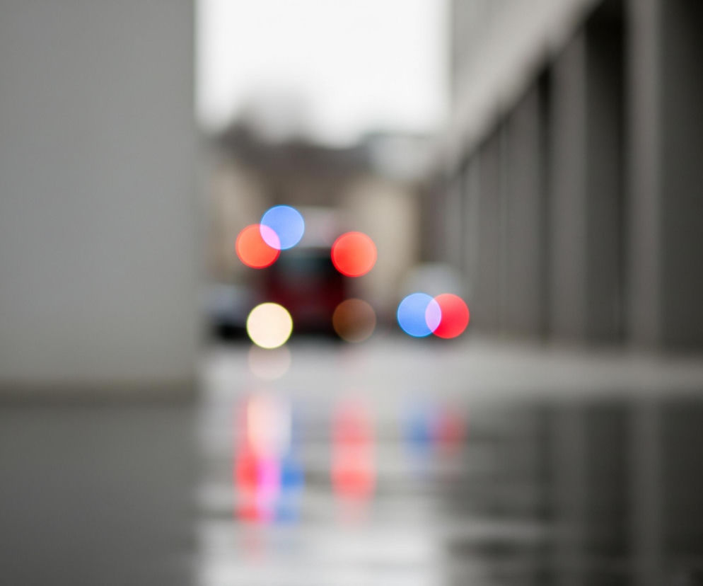 Płytkie pole ostrości rozmywa pierwszy plan i tło, koncentrując uwagę na kolorowych, okrągłych plamach światła w centrum. Lewa strona kadru jest zdominowana przez jasnoszarą, pionową powierzchnię, częściowo odbijającą światło. Po prawej stronie widać szereg ciemniejszych, pionowych kształtów, tworzących kolumnadę lub fasadę budynku, odbijającą się na mokrej, ciemnej powierzchni podłoża. Na linii horyzontu i pod nią widoczne są rozmyte, intensywnie czerwone, niebieskie i żółte plamy świetlne, prawdopodobnie reflektory lub światła sygnalizacyjne pojazdów, które tworzą odbicia w kałużach.