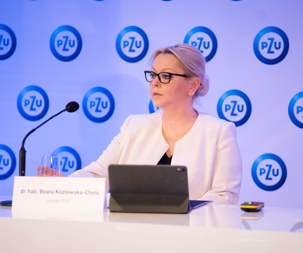 Beata Kozłowska-Chyła, była prezes PZU, w eleganckiej białej marynarce i okularach, siedzi przy stole, obok mikrofonu i szklanki. Tło stanowią powtarzające się loga PZU. Sprawa roszczeń wobec niej to ważny temat, o którym można przeczytać na Super Biznes.