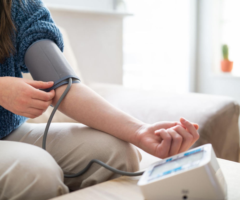 Kobieta w niebieskim swetrze i beżowych spodniach mierzy ciśnienie krwi za pomocą automatycznego aparatu w domowym zaciszu, co jest kluczowe w walce z nadciśnieniem. Więcej o profilaktyce i leczeniu przeczytasz na Poradniku Zdrowie.