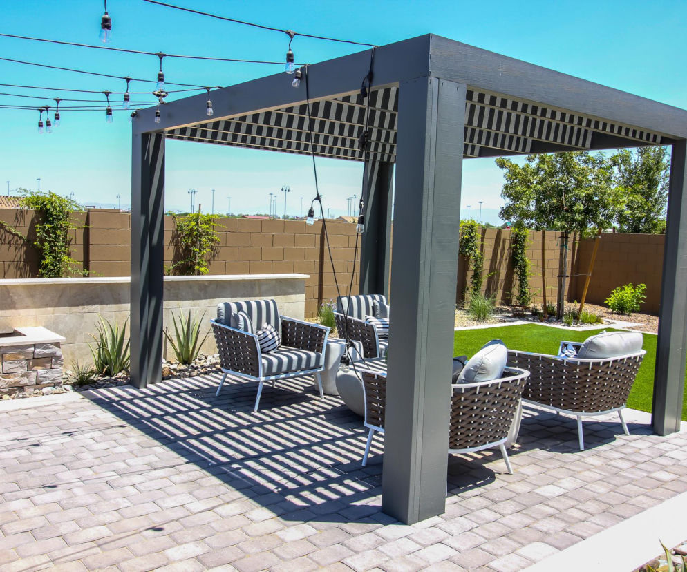Nowoczesna pergola z lamelowym zadaszeniem i ciemnymi ramami, pod którą ustawiono komplet eleganckich, wiklinowych mebli ogrodowych z poduszkami w paski, zapewniająca cień na tarasie wyłożonym kostką brukową. Więcej o aranżacji tarasów przeczytasz na naszym portalu.