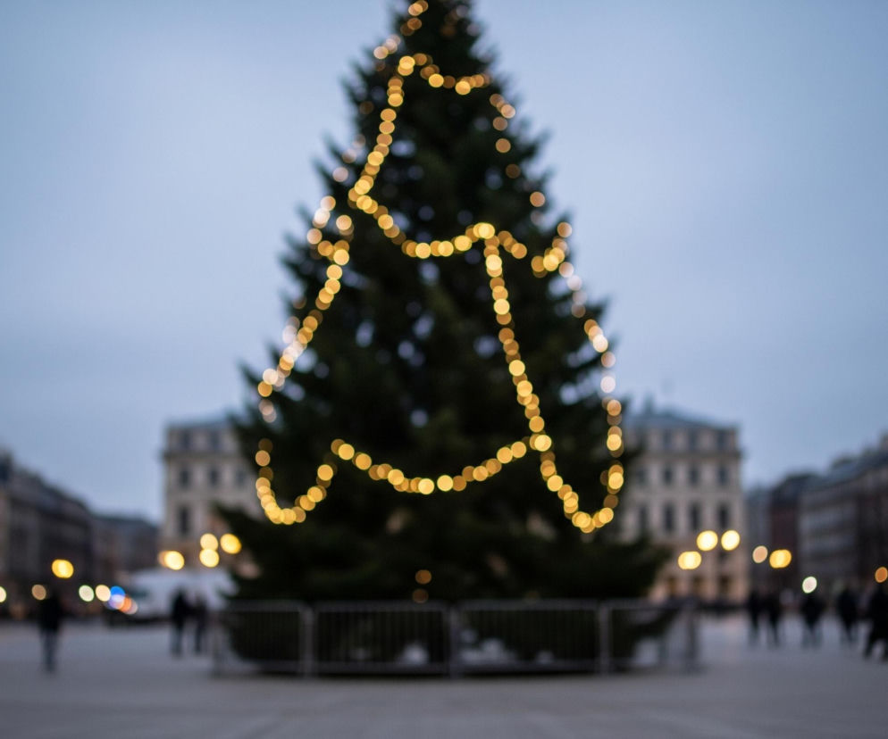 Centralnie na zdjęciu znajduje się duże, zielone drzewo iglaste ozdobione girlandami z ciepłych, żółtych światełek, które są całkowicie rozmyte w efekcie bokeh. Drzewo otacza metalowe ogrodzenie. Tło wypełnia szare, pochmurne niebo oraz rozmyte kontury budynków po obu stronach ulicy, z których niektóre mają włączone światła. Na ulicy widać niewyraźne sylwetki ludzi oraz jaskrawe, okrągłe plamy świateł ulicznych i samochodowych.