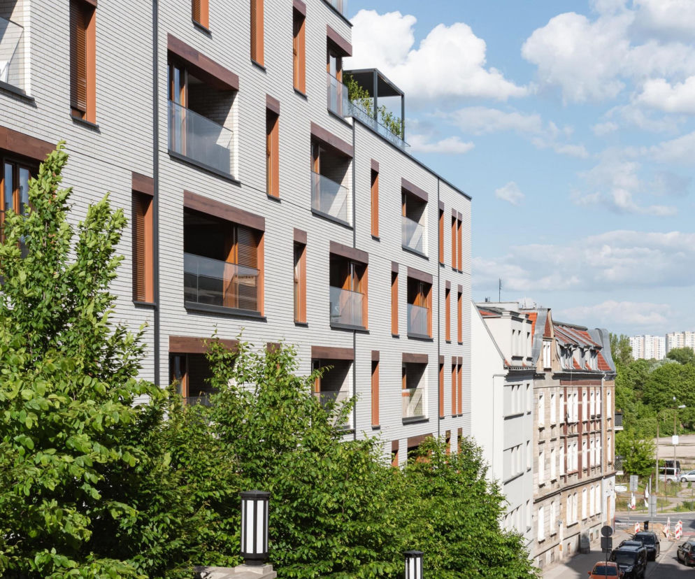 Nowoczesny budynek Vilda Moderne w Poznaniu, z szarymi fasadami i brązowymi detalami okiennymi, harmonijnie wpisuje się w otoczenie. Widoczne są tarasy, zielona roślinność i zabytkowe schody, podkreślające dialog z historyczną tkanką miejską, o czym przeczytasz na Architektura Murator Plus.