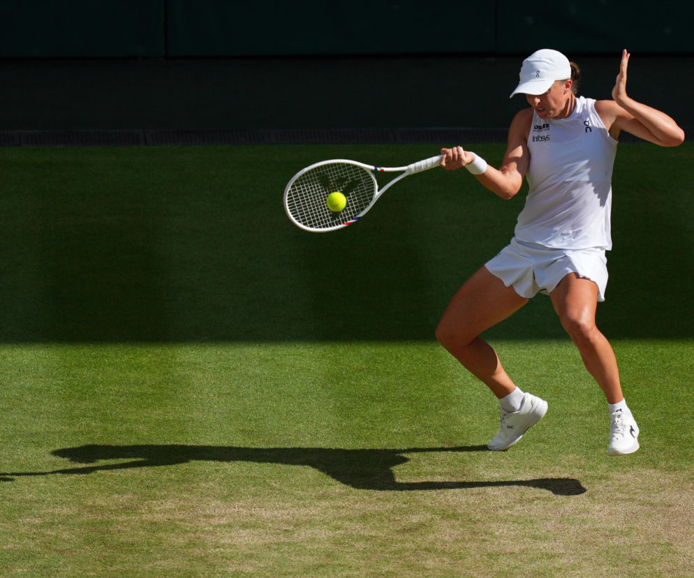 Tenisistka Iga Świątek, ubrana w biały strój sportowy, z rakietą uderza żółtą piłkę na korcie Wimbledonu. O tym, kiedy grają Polacy na Wimbledonie, przeczytasz na naszym portalu.