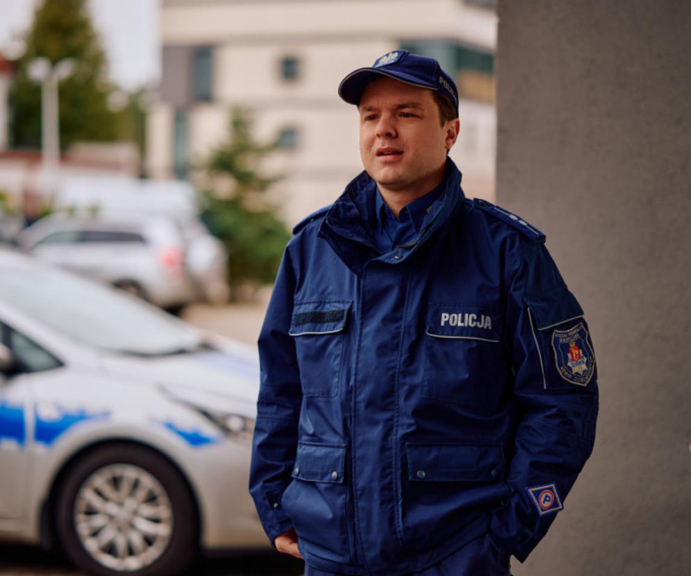 Mężczyzna w policyjnym mundurze i czapce, Maciej Kobiela jako Remek z serialu Na Wspólnej, stoi na tle radiowozu i budynku. W artykule na naszym portalu przeczytasz o jego szokującym odkryciu złodziejek w rodzinie.