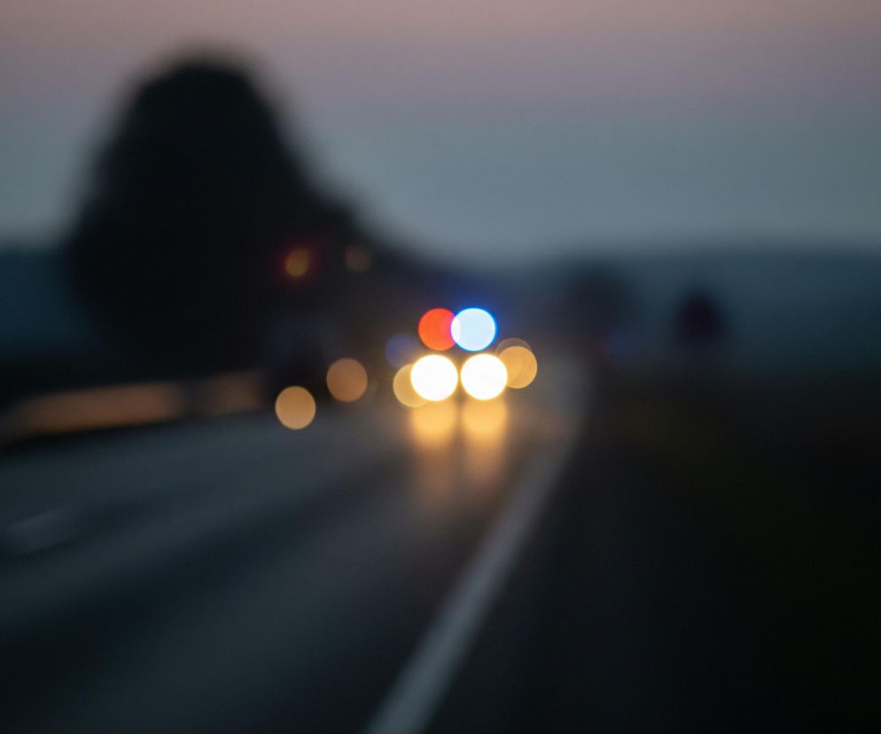 Rozmyty obraz przedstawia nocną drogę o zmierzchu, z jasnymi punktami świateł pojazdów w oddali. Na pierwszym planie widoczny jest ciemny asfalt drogi, z wyraźną białą linią podziału pasa ruchu po prawej stronie, biegnącą ku górze. W oddali, w centrum kadru, znajdują się rozmyte światła pojazdu, składające się z dwóch pomarańczowych i dwóch jasnych, niemal białych kręgów, a nad nimi dwa mniejsze kręgi, jeden czerwony i jeden niebieski. Horyzont ukazuje ciemną, rozmytą sylwetkę drzewa po lewej stronie i delikatny, różowo-szary odcień nieba w górnej części.