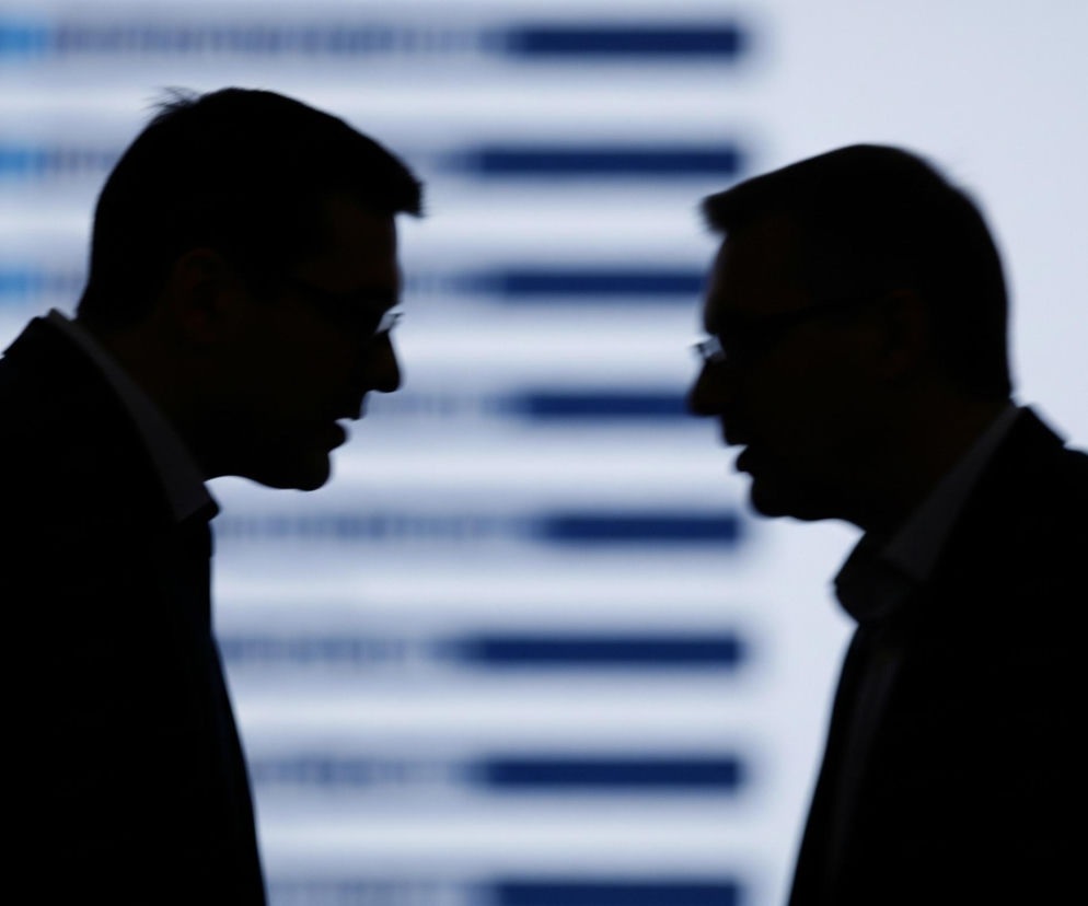 Dwaj mężczyźni stoją przodem do siebie, ich sylwetki są widoczne w ciemnej kontraście na tle jasnego ekranu. Obaj mężczyźni noszą okulary i wydają się być ubrani w koszule z kołnierzykami i marynarki. W tle, rozmyte niebieskie i białe pasy biegną poziomo przez całą szerokość ekranu.