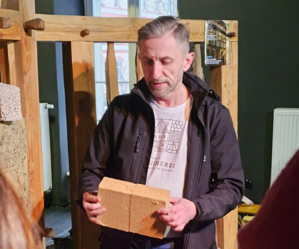Darek Borkowski z Fundacji Ruinersi na Dolnym Śląsku, w czarnej kurtce i białej koszulce z grafiką domów, prezentuje jasny, prostokątny cegłę. Rozmawia o zmianach w myśleniu o remontach starych domów, co szczegółowo opisujemy na naszym portalu.