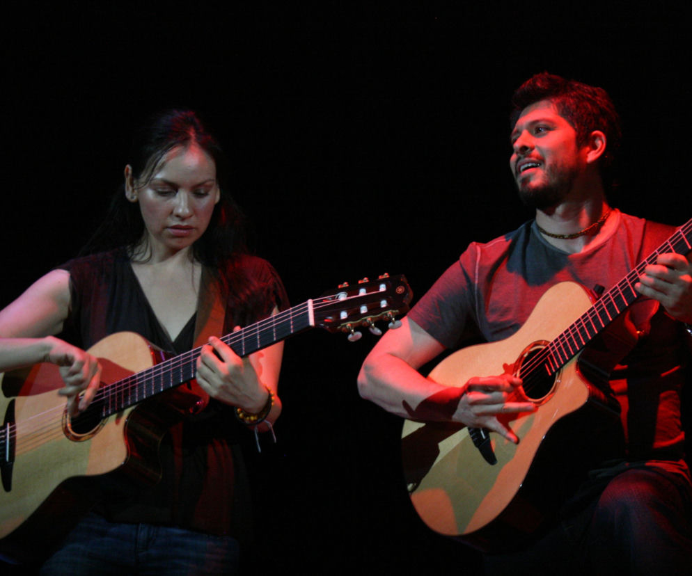 Meksykański duet Rodrigo y Gabriela grający na gitarach akustycznych na ciemnej scenie, oświetlonej czerwonym światłem. Gabriela po lewej skupiona na instrumencie, Rodrigo po prawej uśmiecha się podczas występu. Więcej o koncercie tych wirtuozów na naszym portalu.