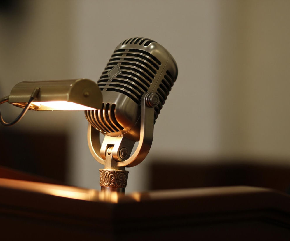 Na pierwszym planie, z lekkim odchyleniem w prawo, widoczny jest metalowy, chromowany mikrofon w stylu retro, umieszczony na zdobionej, złotej podstawie, która spoczywa na drewnianej mównicy. Obok mikrofonu, po jego lewej stronie, znajduje się mała, złota lampka z ruchomym ramieniem, emitująca ciepłe, żółte światło, rozświetlające część mównicy i przód mikrofonu. Tło jest rozmyte, utrzymane w ciepłych odcieniach beżu i brązu, z niewyraźnymi ciemnymi elementami po prawej stronie.