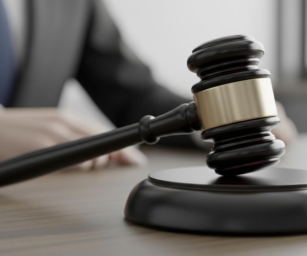 Młotek sędziowski wykonany z ciemnego drewna z złotym pierścieniem na główce leży ukośnie na ciemnej, okrągłej podstawce, umieszczonej na drewnianym blacie. W tle, częściowo rozmyta postać w ciemnym garniturze i niebieskim krawacie siedzi za blatem. Jasne światło wpada z prawej strony, oświetlając scenę.