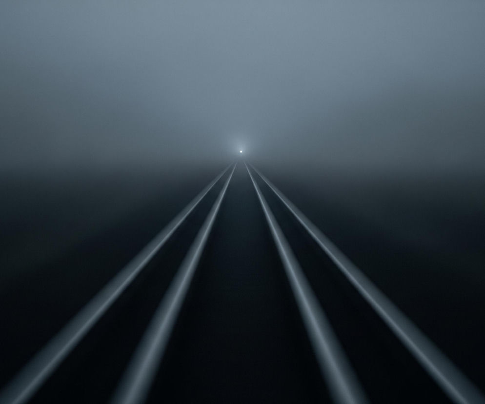 W centrum obrazu widoczne są cztery jasne, równoległe linie, które zbiegają się w oddali, tworząc wrażenie torów lub drogi. Linie te są otoczone przez gęstą, jednobarwną mgłę lub dym, która rozciąga się od ciemnego dołu do jaśniejszej góry. W dalekim punkcie zbiegu linii, w centrum horyzontu, znajduje się mały, intensywny punkt świetlny. Całość utrzymana jest w chłodnej palecie barw, od prawie czarnego dołu po jasnoszary, niemal biały górny obszar, z dominującymi odcieniami szarości i błękitu.