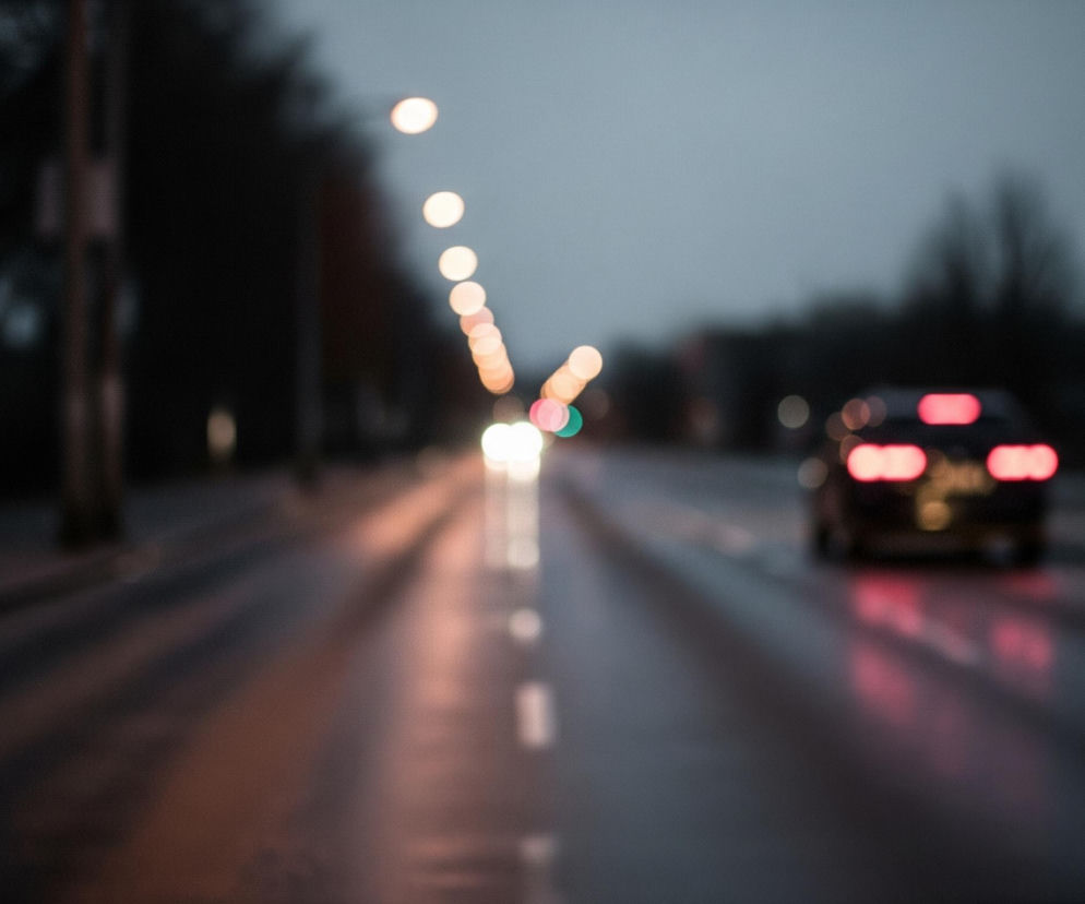 Droga asfaltowa, z widocznym odblaskiem wilgoci i świateł, biegnie przez centrum kadru, rozszerzając się w kierunku przodu. W oddali, po lewej stronie drogi, wznosi się rząd okrągłych, rozmytych świateł ulicznych o ciepłym, pomarańczowym odcieniu, prowadzących perspektywicznie w głąb kadru. Na prawym pasie drogi znajduje się rozmyty samochód z wyraźnie świecącymi, czerwonymi światłami tylnymi. W tle po obu stronach drogi rozpościerają się niewyraźne, ciemne kształty drzew i budynków, a powyżej nich widać pochmurne, szaroniebieskie niebo.