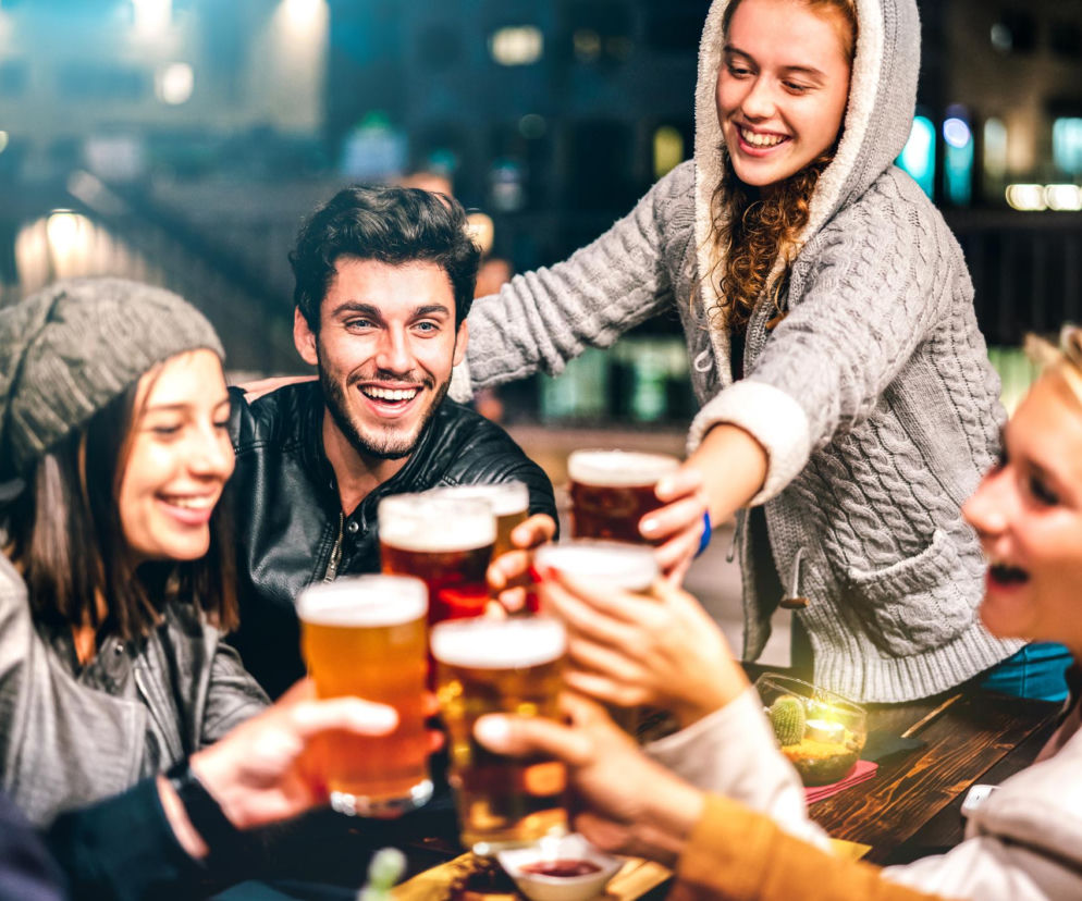 Grupa młodych, roześmianych znajomych wznosi toasty piwem, siedząc przy drewnianym stole w oświetlonym pubie. Artykuł na Poradnik Zdrowie omawia, dlaczego piwo bezglutenowe może nadal powodować objawy podobne do IBS u osób z nadwrażliwością jelitową, analizując obecność fruktanów i białek ATI.