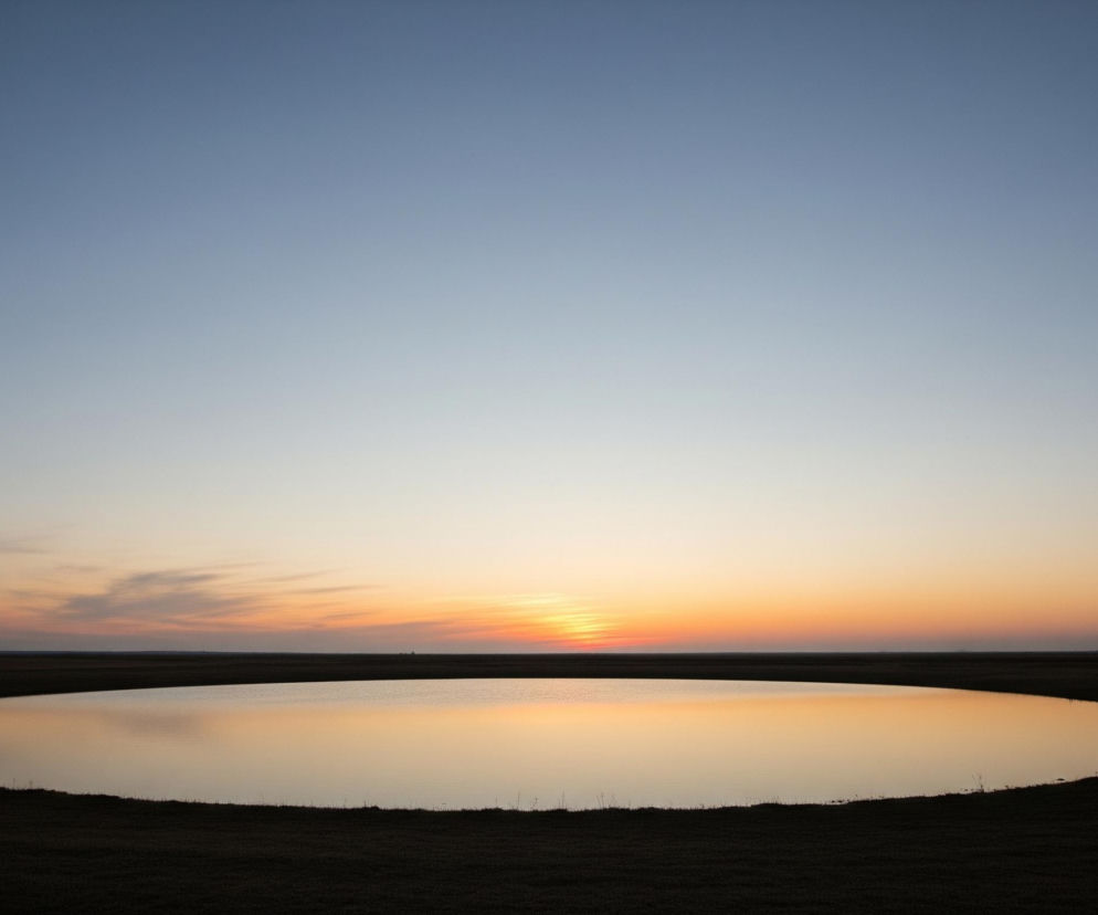 Centralne miejsce w dolnej części kadru zajmuje owalne, płytkie jezioro lub staw, którego powierzchnia odbija jasne, pomarańczowo-żółte światło zachodzącego słońca. Wokół zbiornika wodnego rozciąga się ciemna, niemal czarna równina, prawdopodobnie porośnięta trawą lub niską roślinnością. Horyzont przecina zdjęcie, oddzielając ciemną ziemię od rozległego nieba, które przechodzi od błękitu w górnej części kadru, przez jasne, pomarańczowe i żółte odcienie w centrum, aż do delikatnego różu i błękitu tuż nad linią horyzontu. Po lewej stronie nieba widoczne są subtelne, rozproszone obłoki.