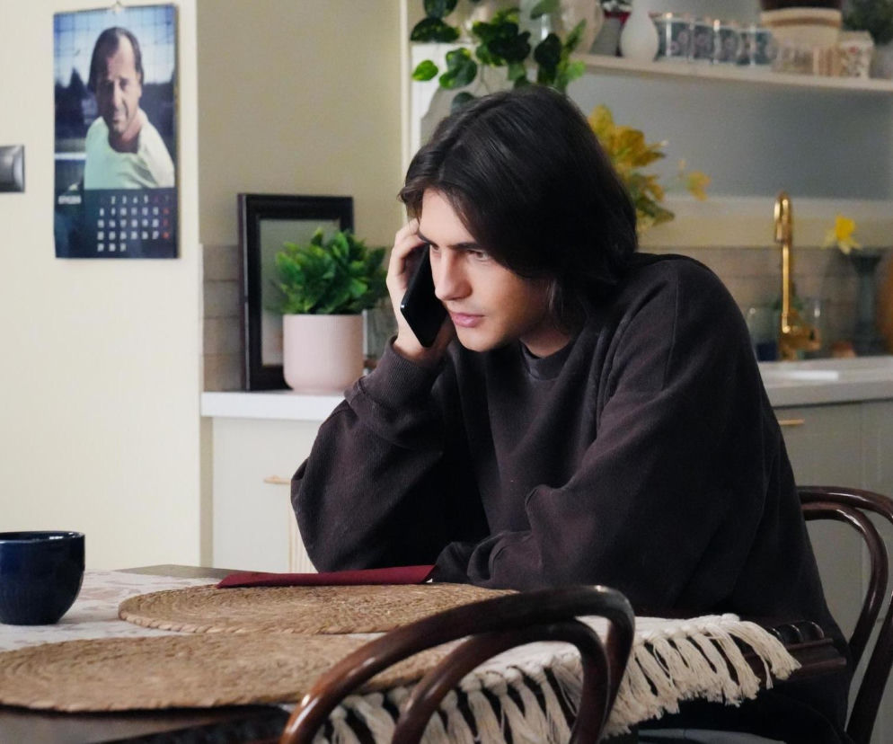 Romeo (Jacek Tyszkiewicz) z serialu Barwy szczęścia w ciemnej bluzie siedzi przy stole, trzymając telefon przy uchu, jego twarz wyraża zaniepokojenie. W tle widoczna kuchnia z białymi szafkami i złotym kranem. Obok na ścianie wisi kalendarz. Więcej o dramatycznym telefonie Romea przeczytasz na Super Seriale.