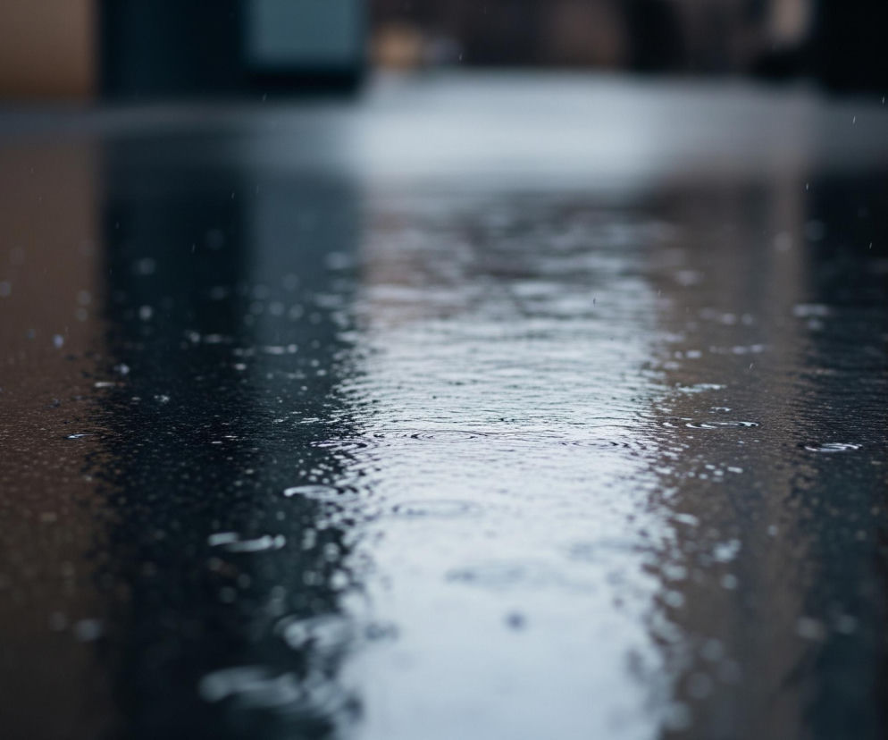 Ciemna, mokra nawierzchnia zajmuje większą część kadru, z wyraźnymi refleksami światła odbijającymi się od jej powierzchni. Widoczne są liczne, drobne krople wody, niektóre z nich tworzą koncentryczne fale na powierzchni, świadcząc o padającym deszczu. W tle, rozmyte i niewyraźne, rysują się ciemniejsze kształty, które mogą być budynkami lub innymi elementami miejskiego krajobrazu, tworząc delikatne, rozmazane odbicia na mokrej nawierzchni.