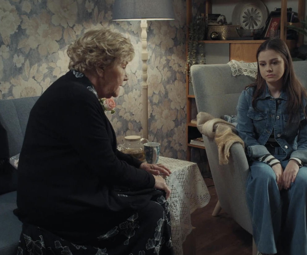 Barbara (Teresa Lipowska) i Basia (Karina Woźniak) siedzą w pokoju. Barbara, ubrana w ciemną bluzkę i spódnicę w białe wzory, z rozłożonymi dłońmi na serwetce przy dzbanku herbaty. Basia w dżinsowej kurtce i spodniach, siedzi na krześle ze spuszczoną głową. Zdjęcie ilustruje scenę ze złamanym sercem w odcinku M jak miłość. Więcej na Super Seriale.