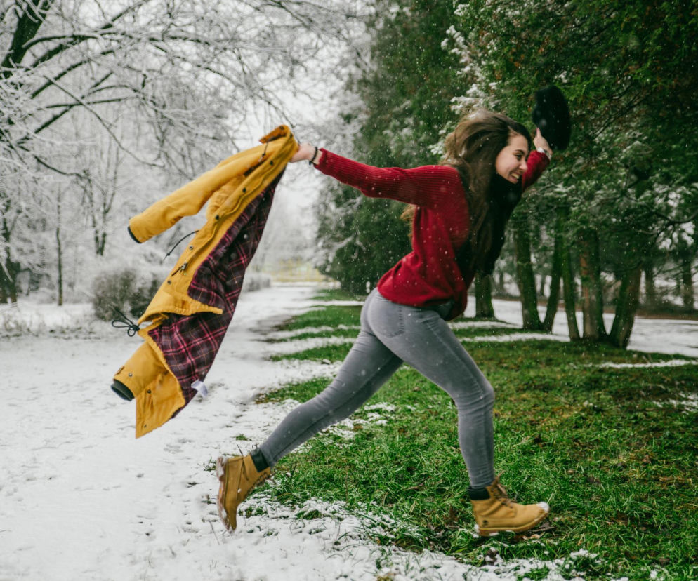 Szczęśliwa kobieta biegnąca ze zdjętą kurtką po pokrytej śniegiem ścieżce i zielonej trawie. Przedwiośnie ze zmieniającą się pogodą może wpływać na samopoczucie. Więcej o wpływie pogody na zdrowie przeczytasz na Poradnik Zdrowie.