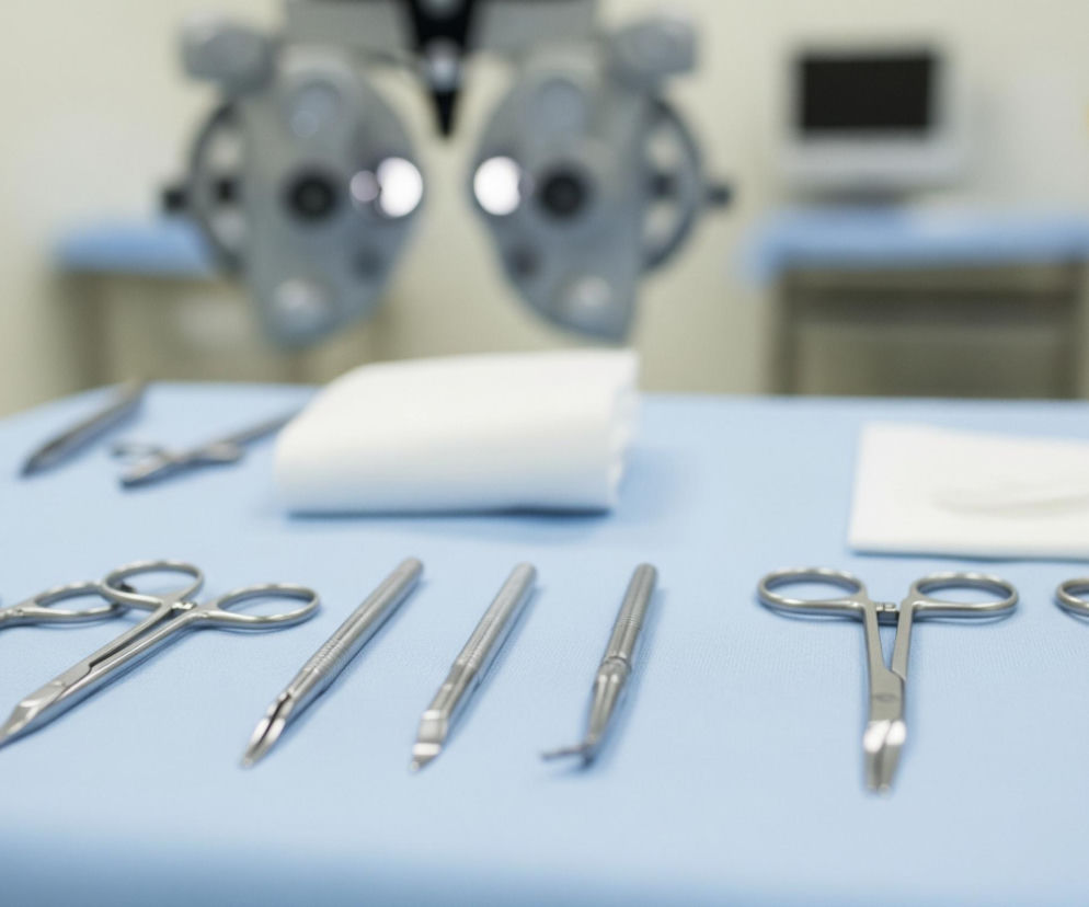 Na jasnoniebieskim, teksturowanym obrusie, leży zestaw metalowych narzędzi chirurgicznych oraz białe, złożone kawałki materiału. Po lewej stronie widoczne są nożyczki o wąskich, spiczastych ostrzach i owalnych uchwytach, obok nich dwa podłużne narzędzia z delikatnie karbowanymi trzonkami i ostrymi końcówkami. Dalej, po prawej stronie, leży podobny zestaw narzędzi, w tym metalowe szczypce z owalnymi uchwytami i krótkimi, spłaszczonymi końcówkami. W tle, częściowo rozmyte, znajdują się fragmenty maszyn i urządzeń, w tym ciemnoszare, podwójne urządzenie optyczne oraz prostokątny monitor z ciemnym ekranem.