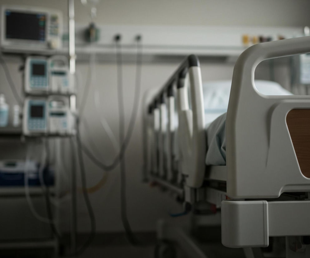 Na pierwszym planie, po prawej stronie, znajduje się jasnoszare, puste łóżko szpitalne z podniesioną częścią zagłówkową. Po lewej stronie łóżka widoczne są szare barierki, a jego boki wypełniają prostokątne panele imitujące drewno w kolorze jasnobrązowym. W tle, po lewej stronie obrazu, stoją pionowo ułożone urządzenia medyczne z ekranami monitorów, podłączone do szarej ściany za pomocą licznych przewodów. Na samej górze ściany przebiega pozioma, szara listwa techniczna.