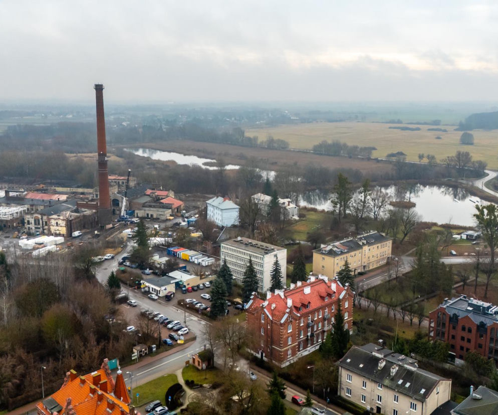 Widok z lotu ptaka na Królewską Papiernię w Konstancinie-Jeziornie, z charakterystycznym wysokim kominem dominującym nad terenem przemysłowym, otoczonym zielenią i zbiornikami wodnymi, co nawiązuje do rewitalizacji tego obszaru przez Grupę Arche. Więcej o inwestycji przeczytasz na Architektura Murator Plus.