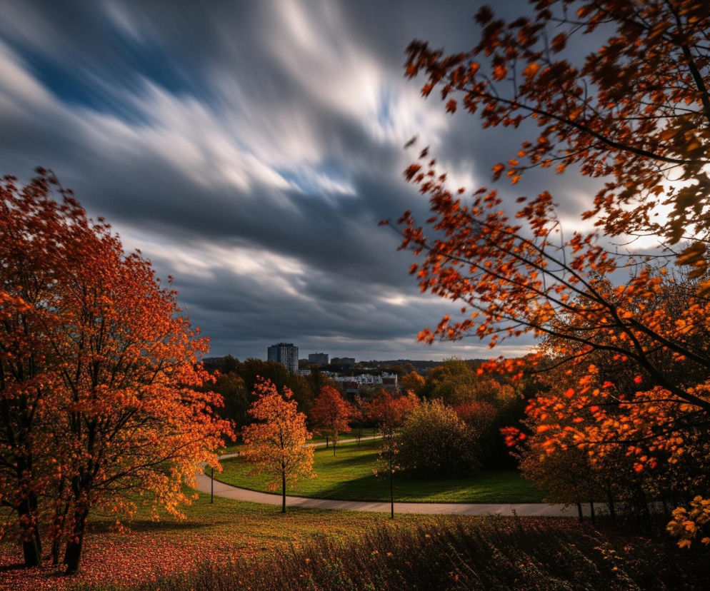 Park w jesiennych barwach, z pomarańczowo-czerwonymi liśćmi drzew na pierwszym planie, rozciągającymi się po bokach i na dole kadru. Centralną część zajmuje zielona trawa, przez którą wije się jasna, utwardzona ścieżka z kilkoma lampami parkowymi. W tle, za drzewami, majaczą budynki miejskiej zabudowy pod dynamicznym niebem z rozmytymi, smugowymi chmurami, które zdają się przesuwać z dużą prędkością. Słońce, częściowo przesłonięte przez gałęzie i liście, tworzy jasną poświatę w prawym górnym rogu.
