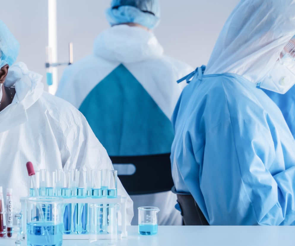 Naukowcy w kombinezonach ochronnych pracujący w laboratorium, jeden z nich korzysta z mikroskopu, a na stole widoczne są probówki z niebieskimi i czerwonymi płynami. W kontekście lekooporności bakterii, o której przeczytasz na Poradnik Zdrowie, obraz symbolizuje intensywne badania nad nowymi rozwiązaniami w medycynie.