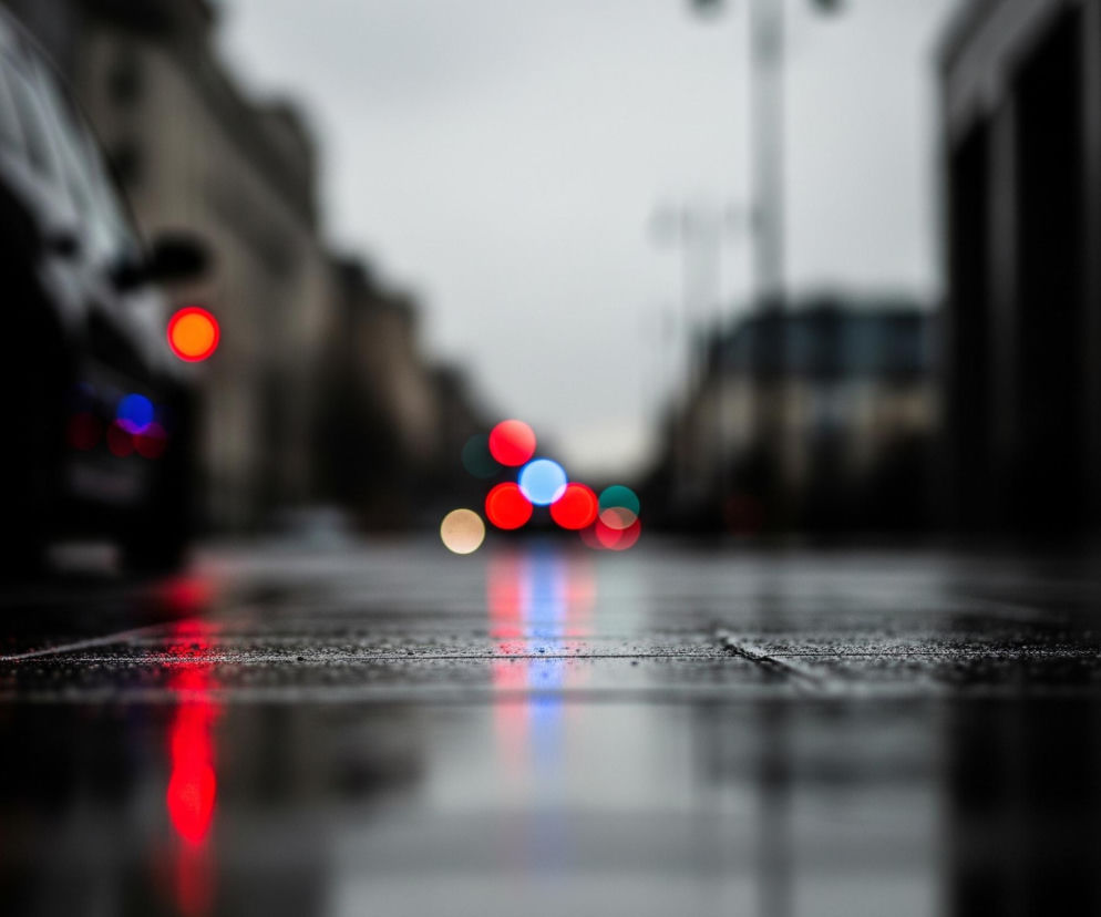 Mokrą asfaltową drogę o zmierzchu lub w nocy, z ostrością ustawioną na drobne krople wody na powierzchni jezdni. Na pierwszym planie, w lewym dolnym rogu, znajduje się czarny fragment samochodu z rozmazanym czerwonym światłem pozycyjnym, a pod nim widać rozmazane niebieskie światło. W tle, powyżej drogi, widoczne są rozmyte światła samochodów i budynków, tworzące kolorowe kręgi bokeh w odcieniach czerwieni, błękitu, pomarańczu i turkusu. Czerwono-niebieskie odbicia świateł odbijają się od mokrej nawierzchni, tworząc podłużne smugi. Górna część zdjęcia jest w dużej mierze rozmyta, ukazując niewyraźne zarysy miejskiego krajobrazu pod szarym niebem.