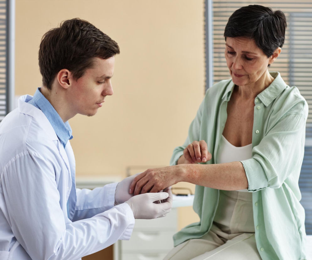 Lekarz w białym kitlu i jednorazowych rękawiczkach delikatnie bada skórę na przedramieniu starszej pacjentki, która ma na rękach czerwone pręgi. O dermografizmie i jego objawach przeczytasz na Poradnik Zdrowie.