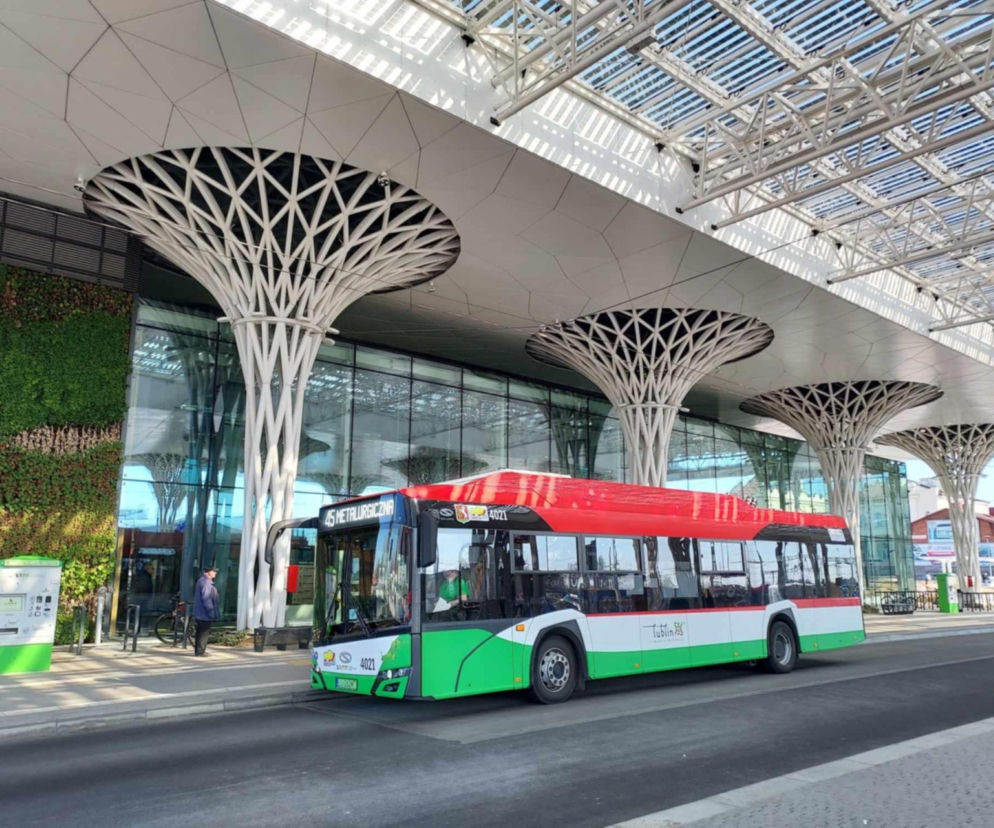 Autobus komunikacji miejskiej w Lublinie, w barwach miasta (czerwono-zielono-białych), stoi pod nowoczesnym przystankiem z dekoracyjnymi filarami. Zdjęcie ilustruje wakacyjne zmiany w rozkładzie jazdy, o których można przeczytać na naszym portalu.
