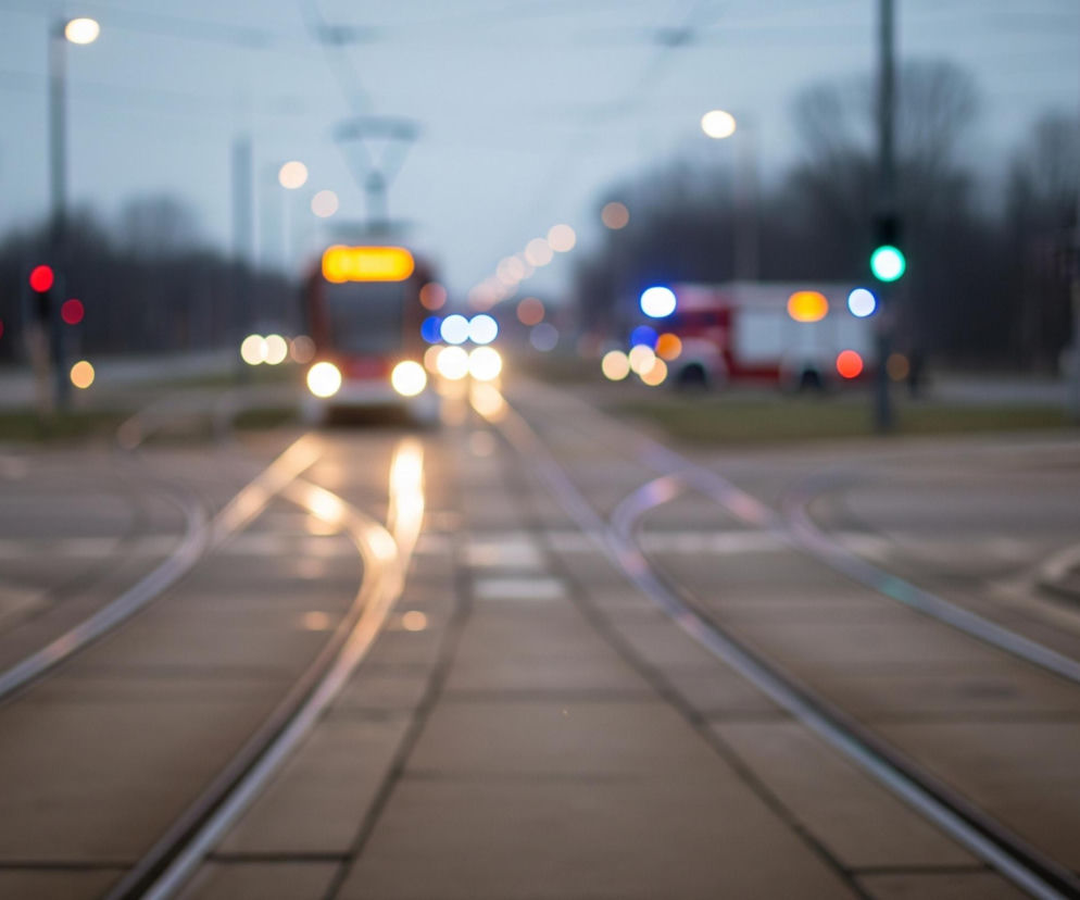 Torowisko tramwajowe z rozjazdami dominuje na pierwszym planie, biegnąc przez środek kadru. W oddali widoczny jest rozmyty czerwony tramwaj z jaskrawymi żółtymi i białymi światłami. Po prawej stronie widać rozmazany pojazd uprzywilejowany z migającymi niebieskimi i czerwonymi światłami, obok zielonego światła sygnalizacji. Cały obraz jest poza ostrością, co tworzy efekt bokeh z licznymi kolorowymi, kolistymi plamami światła na tle ciemnego nieba i niewyraźnych drzew.