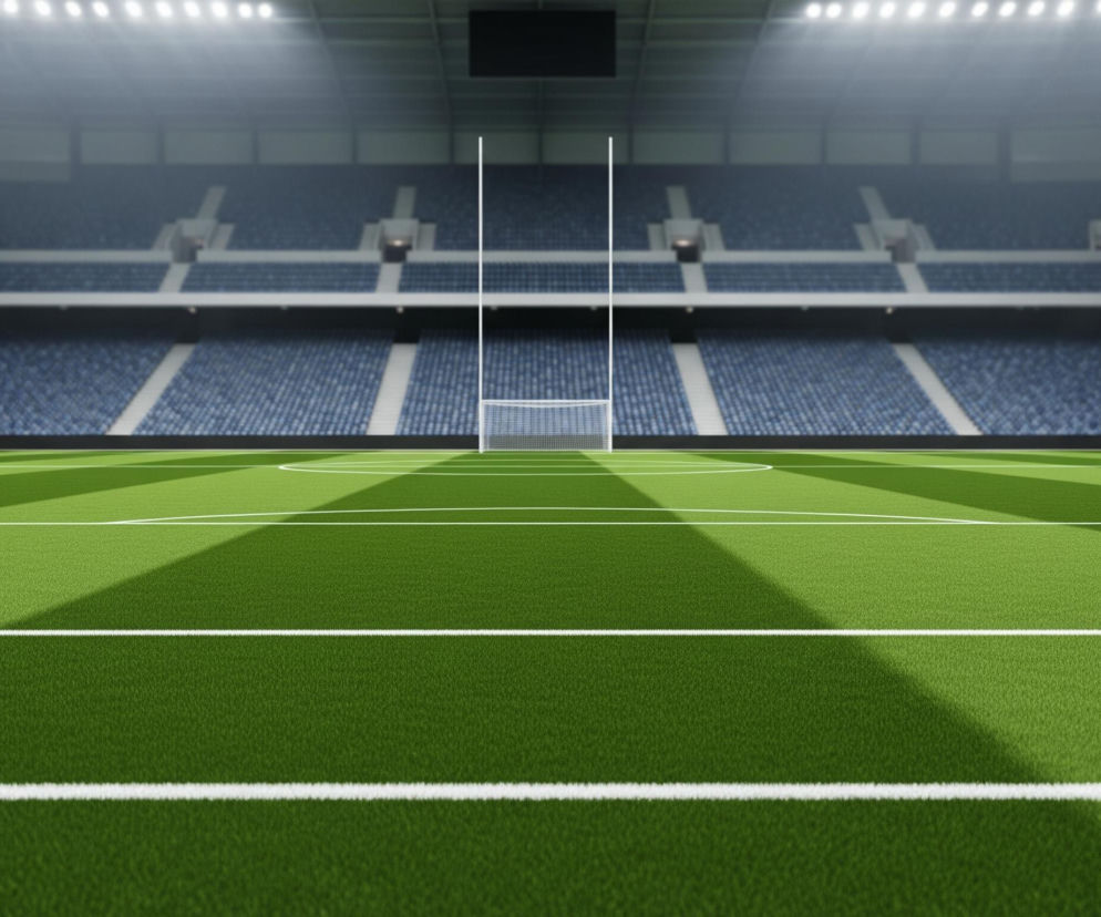 Oświetlony stadion sportowy o zmierzchu z zieloną murawą na pierwszym planie, ozdobioną białymi liniami, które dzielą ją na pasy i okręgi. Na środku boiska znajduje się biała bramka z siatką, a po obu stronach bramki wznoszą się trzy wysokie, białe słupy, z których środkowe są wyższe. Trybuny stadionu, wypełnione rzędami pustych niebieskich siedzeń, rozciągają się w tle, a sufit areny jest widoczny z jasnymi światłami reflektorów.