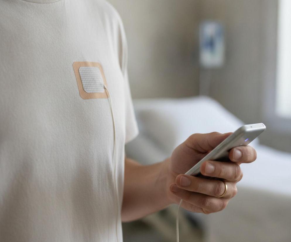 Osoba z przyklejonym ultradźwiękowym rozrusznikiem serca na klatce piersiowej pod białą koszulką trzyma smartfon. To innowacyjne rozwiązanie, o którym przeczytasz na portalu Poradnik Zdrowie, może zastąpić tradycyjne implanty kardiologiczne.