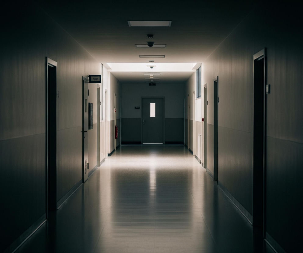 Długi, prosty korytarz jest oświetlony odległym, centralnym źródłem światła, tworząc silną perspektywę i kontrast między jasnym środkiem a ciemnymi krawędziami. Ściany boczne, drzwi i sufit wpadają w ciemność, podczas gdy podłoga odbija światło, tworząc lśniącą smugę. W głębi korytarza widać szare drzwi z pionowym oknem i wiszącymi tabliczkami informacyjnymi po lewej stronie. Na suficie rozmieszczone są prostokątne lampy fluorescencyjne oraz kilka kamer monitoringu.