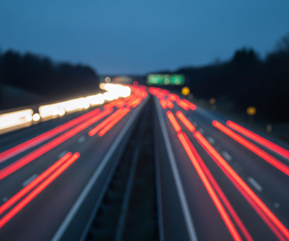Autostradę z smugami świateł pojazdów w nocy, z efektem długiej ekspozycji. Lewa strona jezdni wypełniona jest poziomymi, rozmazanymi smugami białego i jasnożółtego światła, podczas gdy prawa strona ukazuje liczne, czerwone smugi światła, biegnące równolegle. Droga ma po trzy pasy ruchu w każdym kierunku, oddzielone pasem zieleni z barierami, a w oddali majaczy zielony znak drogowy i żółte migające światła. Horyzont tworzy ciemne niebo i zamazany las po obu stronach autostrady.