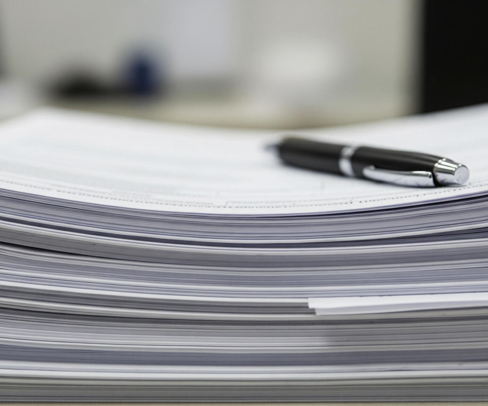 Na pierwszym planie widoczny jest stos dokumentów wydrukowanych na białych kartkach, lekko pofalowanych na krawędziach, tworzących wiele warstw. Na wierzchu sterty leży długopis z czarnym korpusem i srebrnymi elementami, skierowany w prawą stronę. Tło jest rozmyte, ukazując nieostre obszary w odcieniach szarości i bieli, z ciemniejszymi plamami sugerującymi inne przedmioty biurowe.