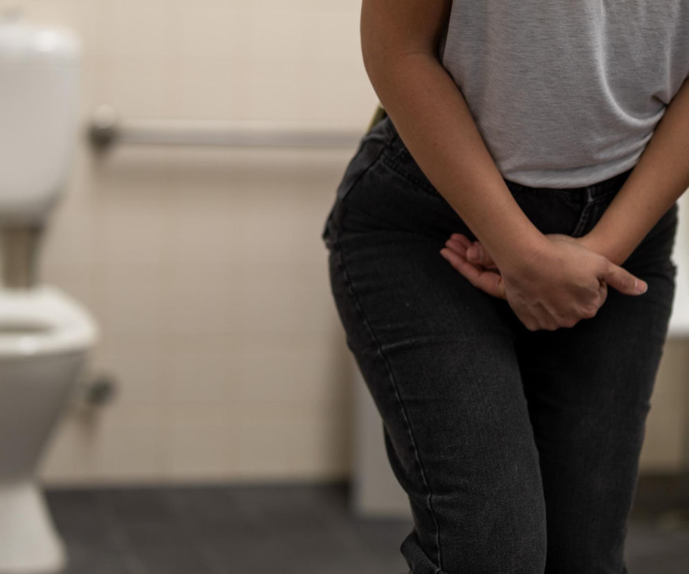 Kobieta z rękami skrzyżowanymi na podbrzuszu, stojąca w łazience z toaletą w tle, symbolizuje nagłe parcie na pęcherz, typowe w stresie lub na mrozie. Na Poradnik Zdrowie znajdziesz informacje, kiedy częste oddawanie moczu jest naturalne, a kiedy świadczy o nerwicy pęcherza.