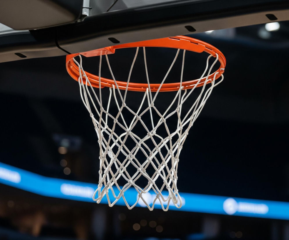 Centralnym elementem obrazu jest kosz do koszykówki, z jaskrawopomarańczową obręczą i białą siatką. Obręcz jest przymocowana do ciemnego elementu konstrukcyjnego, a powyżej widoczne są metalowe wsporniki i śruby. Tło jest ciemne i rozmyte, z widocznym błękitnym pasem, prawdopodobnie częścią tablicy wyników lub bandy LED, oraz kilkoma rozproszonymi, niewyraźnymi światłami.