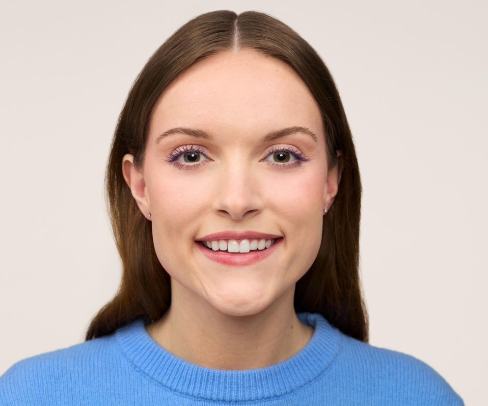 Kobieta z długimi brązowymi włosami i makijażem oczu w odcieniu fioletu, uśmiecha się szeroko. Ubrana w niebieski sweter, prezentuje nowości Benefit Cosmetics, które odmienią makijaż oczu. Więcej o trendach kosmetycznych przeczytasz na naszym portalu.