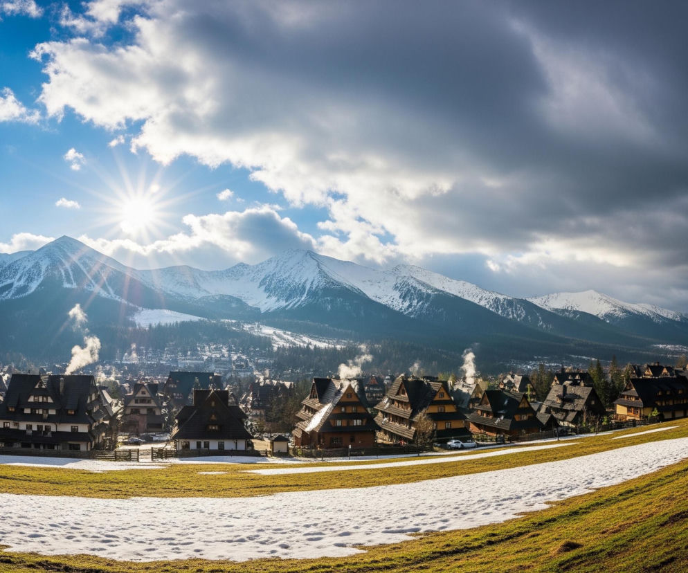Rozległy krajobraz przedstawia pasmo ośnieżonych gór pod częściowo zachmurzonym niebem, z wyraźnym słońcem świecącym po lewej stronie. Na pierwszym planie rozciąga się zielono-żółte zbocze pokryte nieregularnymi płatami białego śniegu, z widocznym ogrodzeniem z drewnianych bali biegnącym wzdłuż zbocza. Poniżej gór leży miasteczko z licznymi drewnianymi domami o stromych, ciemnych dachach, z niektórych kominów unosi się biały dym. W tle za miasteczkiem widoczne są gęste, ciemnozielone lasy, a w oddali inne ośnieżone szczyty górskie.