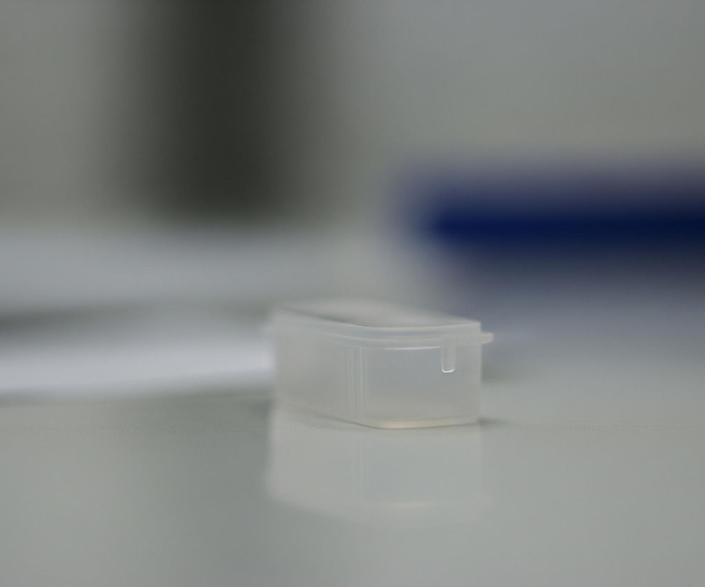 Na pierwszym planie, centralnie, stoi małe, prostokątne pudełko wykonane z półprzezroczystego, białego plastiku. Ma ono gładką powierzchnię i delikatnie odbija światło od spodu, tworząc rozmyte odbicie na jasnej, płaskiej powierzchni, na której stoi. Po prawej stronie pudełka widać wystający, mały, prostokątny uchwyt. Tło jest całkowicie rozmyte, z widocznymi jasnoszarymi obszarami po lewej i ciemniejszym, niebieskim kształtem po prawej, sugerującym jakiś obiekt w oddali.