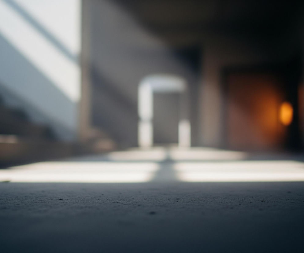 Pofalowana, szara posadzka w pierwszym planie, z drobnymi teksturalnymi nierównościami, rozciąga się w głąb obrazu. Na niej i na ścianie po lewej widoczne są jasne, białe smugi światła, biegnące ukośnie i poziomo, tworząc kontrast z ciemniejszymi partiami pomieszczenia. W oddali, w centrum kadru, znajduje się niewyraźny korytarz z kilkoma przejściami, a na prawo od niego widać rozmyte, ciepłe, pomarańczowe światło. Cały obraz utrzymany jest w stonowanej kolorystyce szarości, bieli i odcieni pomarańczu, z dominującym efektem rozmycia tła.