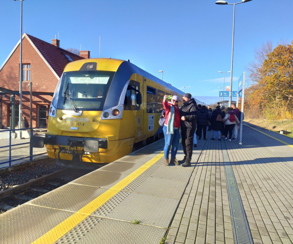 Żółty pociąg na stacji kolejowej w Jastarni, z parą robiącą sobie selfie na peronie i grupą pasażerów czekających na transport. W tle widoczny dworzec oraz jesienne drzewa, symbolizujące modernizację trasy kolejowej Puck-Hel, o której przeczytasz na Super Biznes.